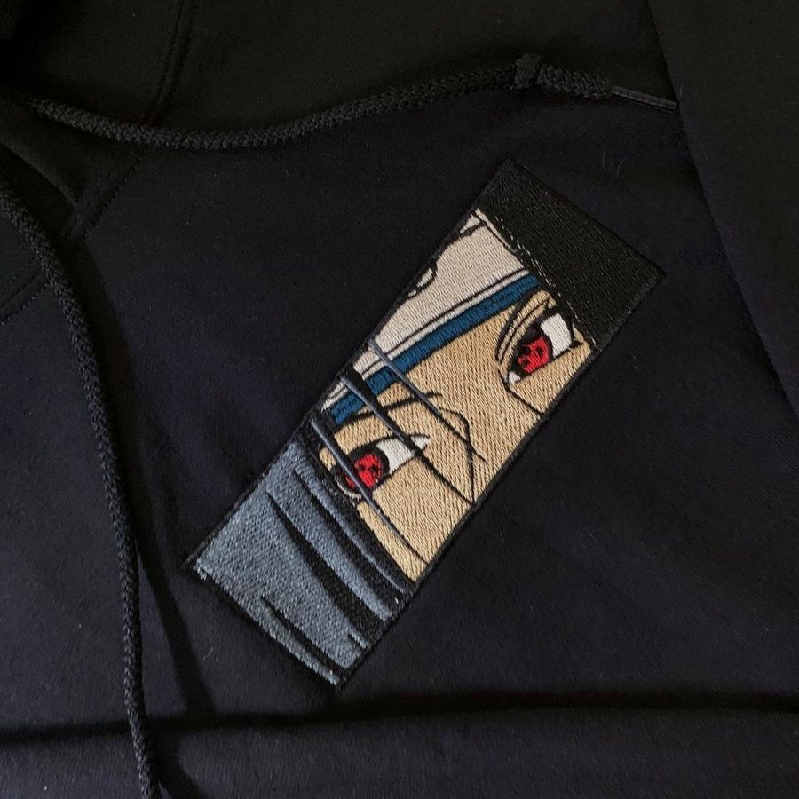 Rectangular embroidered eye panel stitched onto a black hoodie, beside the hood seam and drawstring