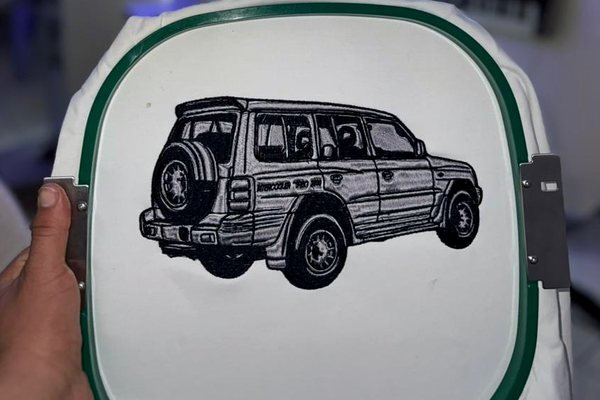 A white panel held in a green embroidery hoop on the machine, embroidered with a detailed line drawing of a four-wheel-drive vehicle