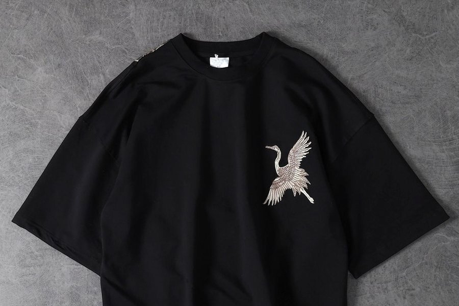 Black oversized t-shirt laid flat on grey concrete, with a small white crane in flight embroidered on the chest