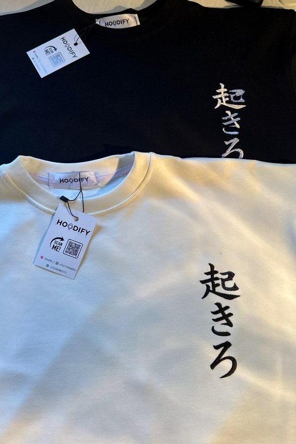 Two folded t-shirts, one black and one bone, both embroidered with the same Japanese script on the chest, Hoodify neck labels and hangtags visible